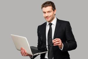 How may I help you Confident young man in formalwear working on laptop while standing against grey background photo