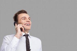 How can I help you Cheerful young man in shirt and tie talking on the phone and smiling while standing against grey background photo