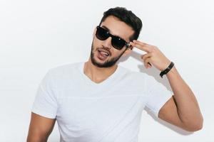 Bang Playful young Indian man in sunglasses gesturing handgun near head and looking at camera while standing against white background photo