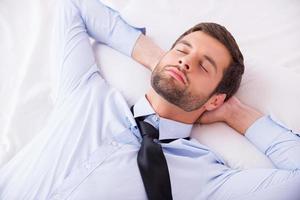 Businessman sleeping. Top view of handsome young man in shirt and tie holding hands behind head while sleeping in bed photo