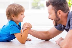 Little champion in arm wrestling. Side view of happy father and son competing in arm wrestling while both lying on the hardwood floor photo