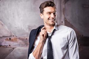 Relaxed handsome. Handsome young man in shirt and tie carrying his jacket on shoulder and looking away with smile while standing against metal background photo