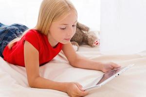 Playing on digital tablet. Cute little girl holding digital tablet and looking at it while lying in bed photo