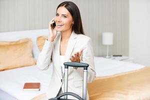 tomando tiempo para llamarla más querida. bella joven mujer de negocios con traje hablando por teléfono móvil y sonriendo mientras se sienta en la cama en la habitación del hotel foto