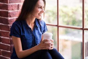Lost in nice thoughts. Cheerful young woman holding cup of hot drink and looking through window while sitting on the window sill photo