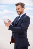 Surfing web in desert. Confident young man in formalwear working on digital tablet and smiling while standing in desert photo