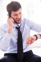 Businessman in a hurry. Furious young man in shirt and tie checking the time and talking on the mobile phone while sitting on the bed photo