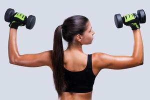 Keeping her body fit and toned. Rear view of young sporty woman exercising with dumbbells while standing against grey background photo