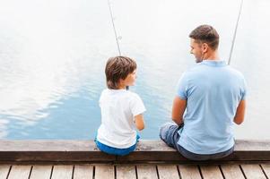 nos encanta pescar juntos. vista trasera de padre e hijo pescando mientras están sentados juntos en el muelle foto