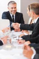 Welcome on board Confident mature man in formalwear shaking hand to one of his colleagues and smiling while sitting at the table together photo