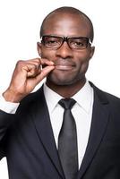 Forced smile. Serious young African man in formalwear closing his mouth by fingers while standing against grey background photo