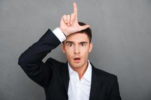 Loser Frustrated young man in formalwear gesturing loser sign and looking at camera while standing against grey background photo