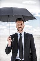 Businessman with umbrella. Confident young man in formalwear holding umbrella overhead while standing in desert photo