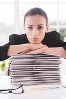 Feeling exhausted. Depressed young woman in formalwear looking at camera and leaning her face at the stack of documents laying on the table photo
