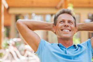 Happy day dreamer. Relaxed mature man holding hands behind head and smiling while outdoors with house in the background photo