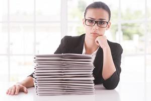 Missing deadlines. Depressed young woman in suit looking at camera and holding head on chin while sitting at the table with stack of documents laying on it photo