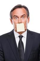 Things to do. Portrait of frustrated mature man in formalwear and adhesive note on his mouth standing against white background and looking at camera photo