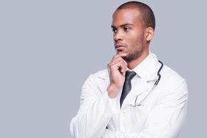 Thoughtful doctor. Thoughtful African doctor holding hand on chin and looking away while standing against grey background photo