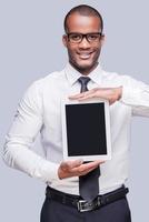 Copy space on his digital tablet. Confident young African man in shirt and tie showing his digital tablet and smiling while standing against grey background photo