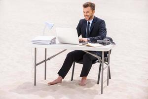 Far away from office. Handsome young man in formalwear working on laptop and smiling while sitting at the table on sand photo