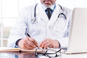 Confident doctor at work. Close-up of mature doctor writing something in note pad while sitting at his working place photo