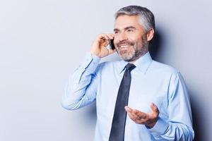 Sharing good news. Happy mature man in shirt and tie talking on the mobile phone and gesturing while standing against grey background photo