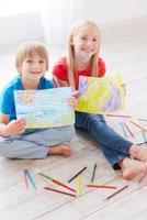 pequeños artistas. dos lindos niños pequeños que muestran las imágenes que dibujan mientras están sentados en el piso de madera dura foto