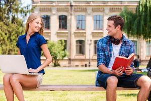 Meeting each other at the first time. Handsome young man sitting on the bench and reading book while beautiful woman sitting near him and using computer photo