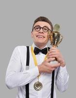 I am the best Cheerful young man in bow tie holding a trophy and looking up while standing against grey background photo