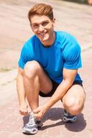 Getting ready to jogging. Handsome man tying shoelaces on sports shoe and smiling while standing outdoors photo