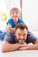 Having fun together. Happy little boy lying on the back of his father and smiling while spending time at home together photo