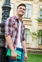 Confident in his bright future. Low angle view of handsome male student holding book and looking up with smile while standing against university building photo