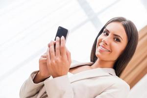 Typing business message. Low angle view of happy young businesswoman in suit holding mobile and looking at camera photo