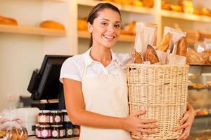 solo pan fresco para nuestros clientes. Hermosa mujer joven en delantal sosteniendo una cesta con pan y sonriendo mientras está de pie en la panadería. foto