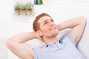 Relaxing at home. Thoughtful young man lying on the couch and keeping hands behind head photo