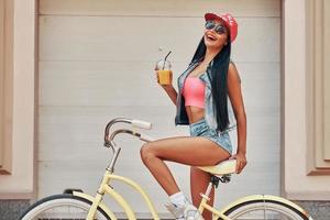 vida jugosa mujer joven alegre sonriendo y sosteniendo un vaso con jugo mientras se sienta en bicicleta contra la puerta del garaje foto