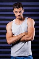 Confident handsome. Sporty young man in tank top keeping arms crossed and looking at camera while standing against striped background photo