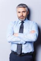 Confident and successful. Confident mature man in shirt and tie looking at camera and keeping arms crossed while standing against grey background photo