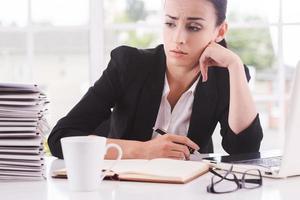 Missing the deadlines. Thoughtful young woman in suit looking at the stack of paperwork and holding head on chin while sitting at her working place photo