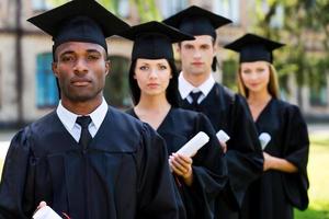 sintiéndose confiado en su futuro. cuatro graduados universitarios de pie en una fila y mirando a la cámara foto