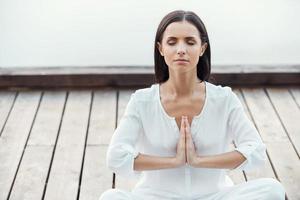 In balance with herself. Beautiful young woman in white clothing sitting in lotus position and keeping hands clasped while meditating outdoors photo