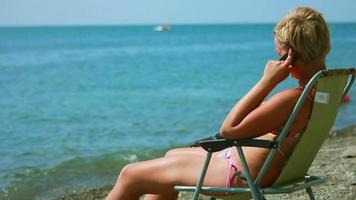 mujer sentada en una tumbona en la playa y hablando por teléfono celular