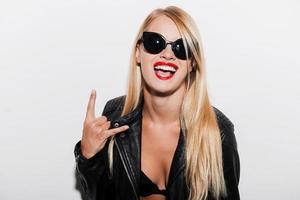 Rock and roll Joyful young woman in black bra and leather jacket looking at camera and gesturing while standing against white background photo