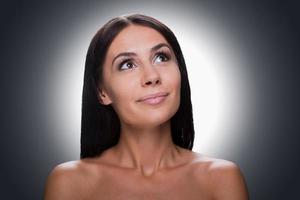 Dreaming of Portrait of thoughtful young shirtless woman looking up and smiling while standing against grey background photo