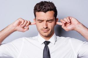 Too loud sound. Frustrated young man in shirt and tie holding fingers in ears and keeping eyes closed while standing against grey background photo