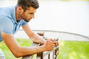 Typing message to friend. Side view of handsome young man typing message on mobile phone while standing on the riverbank photo