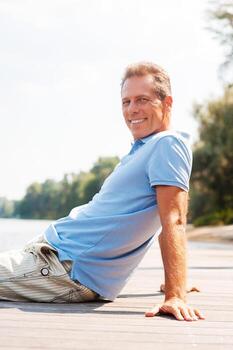 Time to relax. Side view of happy mature man looking away and smiling while sitting at the quayside photo