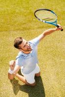 Celebrating success. Top view of happy young man in polo shirt holding tennis racket and gesturing while kneeling on tennis court photo