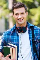 estudiante confiado y exitoso. apuesto joven sosteniendo libros y sonriendo mientras está de pie al aire libre foto