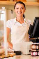 Cheerful cashier. Beautiful young female cashier in apron standing near cash register and smiling photo